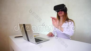 年轻女子坐在桌子旁，拿着银色笔记本电脑，测试虚拟现实眼镜。